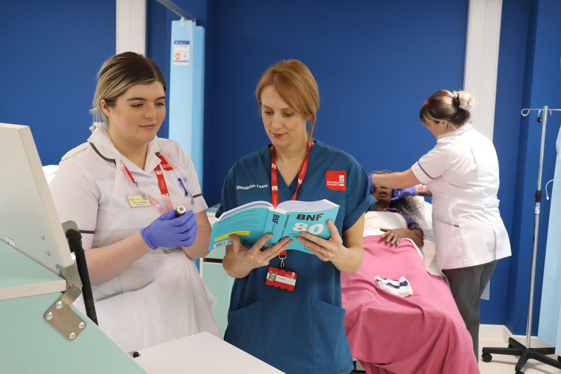A technician looking at a drugs manual with a student, with another student nurse in the background tending to a manikin.